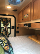 Inside a cozy RV rental: bed with floral pillows, wooden cabinets, window with curtains, and ceiling fan.