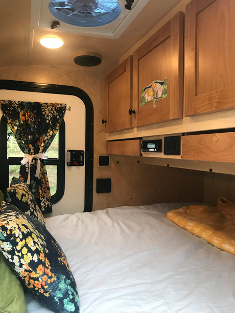 Inside a cozy RV rental: bed with floral pillows, wooden cabinets, window with curtains, and ceiling fan.