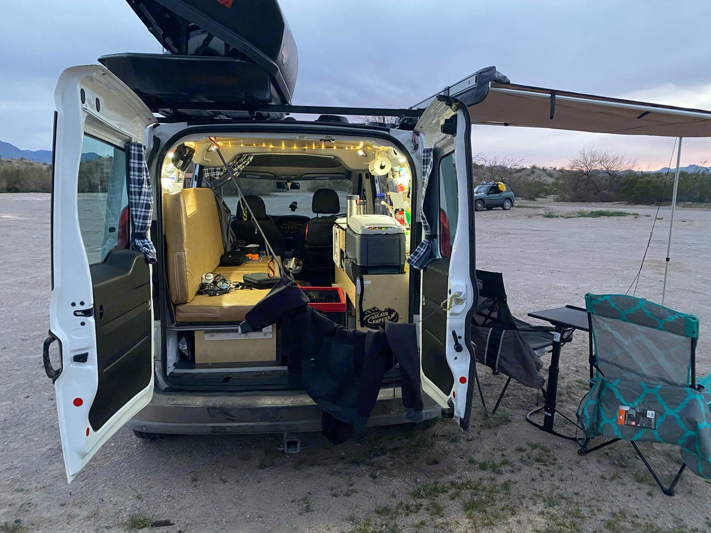 Cozy RV rental: White camper van with open rear doors, lit interior, side awning, and outdoor chairs/table for camping.