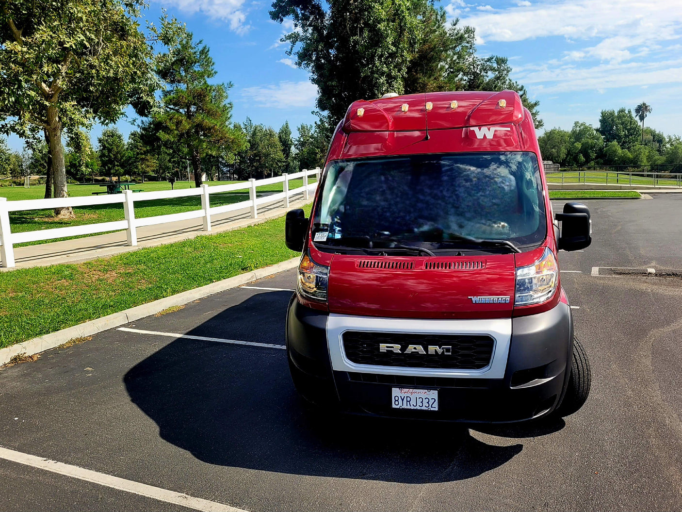 A bright red Winnebago RV rental van is parked on asphalt in a sunny park with green grass and trees.