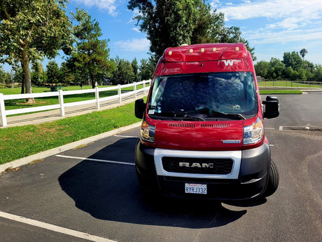 A bright red Winnebago RV rental van is parked on asphalt in a sunny park with green grass and trees.