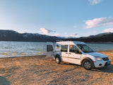 White RV rental van with open door on sandy shore, serene lake & snowy mountains in sunset light.