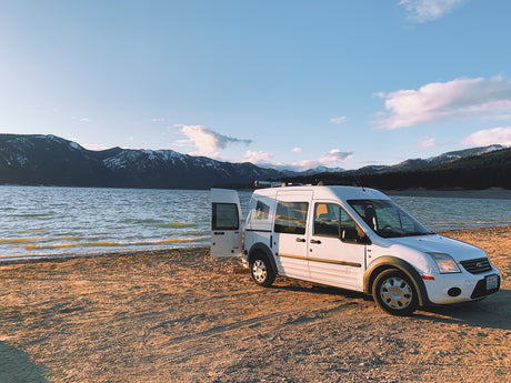 White RV rental van with open door on sandy shore, serene lake & snowy mountains in sunset light.