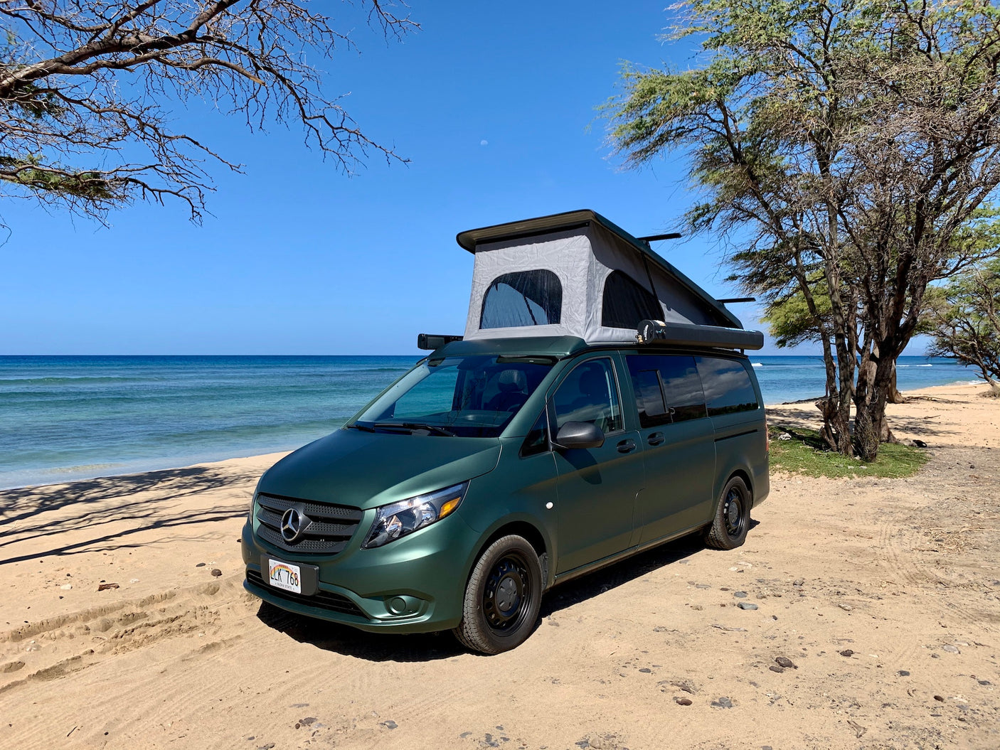 An olive-green Mercedes camper van with a pop-top roof is parked on a sandy beach in Hawaii, perfect for an RV rental.