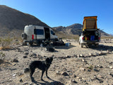Remote camping scene featuring an RV rental van, truck with rooftop tent, and a dog in a rocky desert landscape.