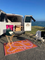 RV rental camper van parked by the ocean with an outdoor cooking setup, chair, and open doors showing the cozy interior.