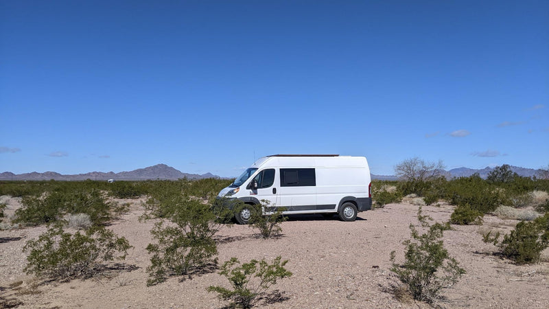 This RV rental camper van is parked amidst desert bushes with mountains in the background under a clear blue sky.|Inside a custom rv rental camper van: wood interior — photo 1