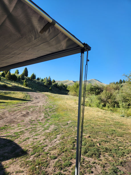 RV rental awning shades a sunny landscape with green hills, trees, and grassy fields under a blue sky.