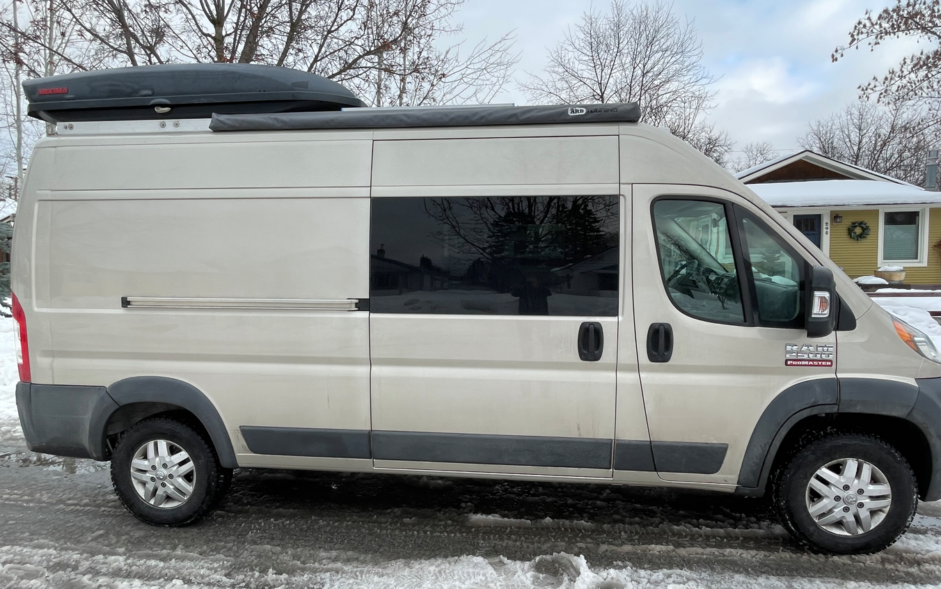 A light-colored RAM ProMaster RV rental is parked on a snowy street, featuring a rooftop cargo box and an awning.