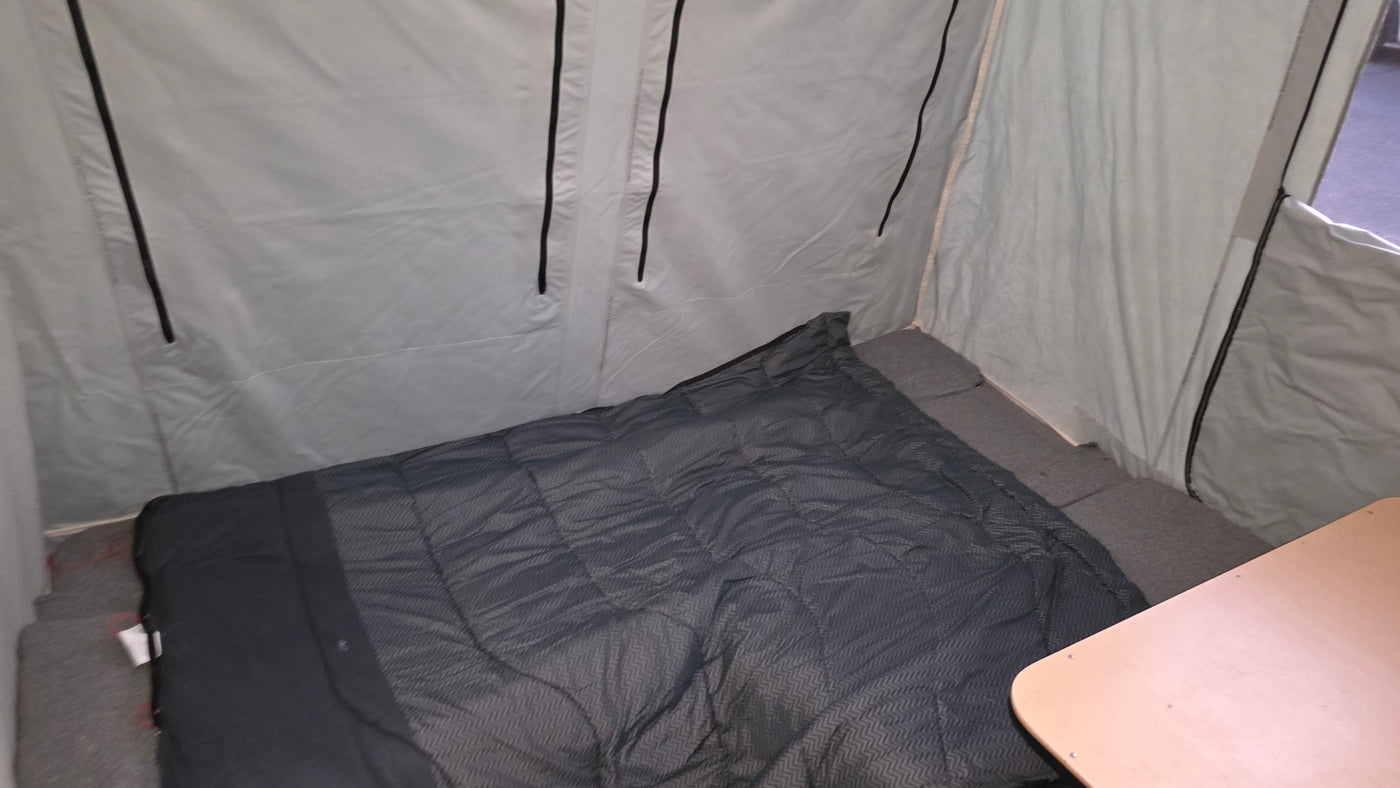 RV rental's compact interior: a dark sleeping bag on a gray bed, light fabric walls, and part of a wooden table. — photo 4