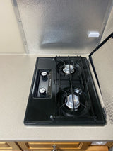 Compact 2-burner gas cooktop in an RV rental kitchen. Features controls, grates, and a hinged cover for mobile cooking.