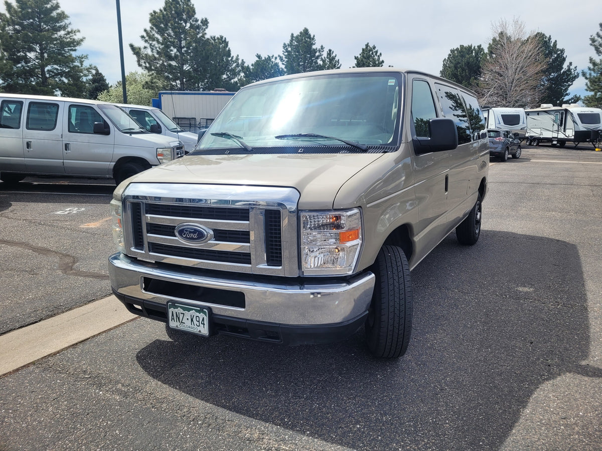 Tan Ford van parked, with other vans & travel trailers/rv rentals in the background. Sunny day in a large lot.