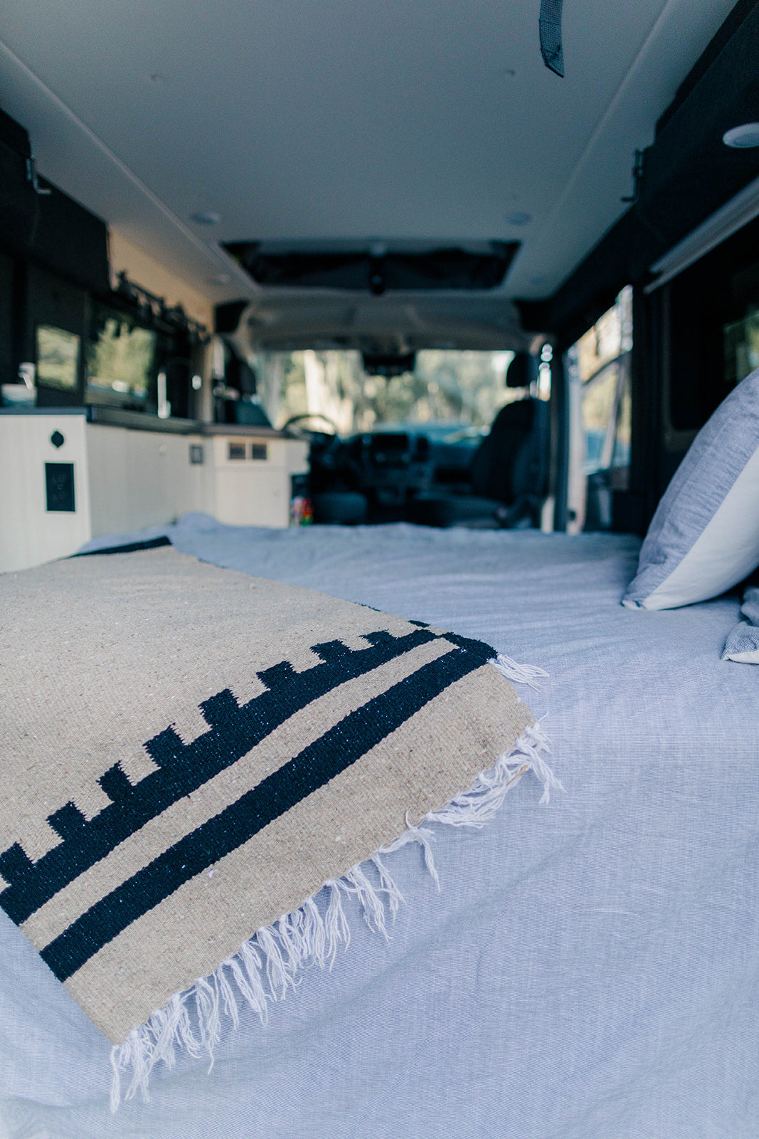 Cozy RV rental interior: a comfortable bed with a patterned blanket fills the foreground, leading to the driver's cabin.