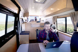 Inside a spacious rv rental, a man works on a laptop at a table, while a woman works on another in the rear bed area. Ideal for remote work.
