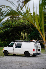White RV rental campervan, ready for tropical adventure. Parked on gravel, framed by palm fronds & lush mountains.