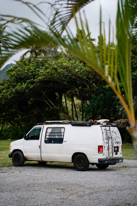 White RV rental campervan, ready for tropical adventure. Parked on gravel, framed by palm fronds & lush mountains.