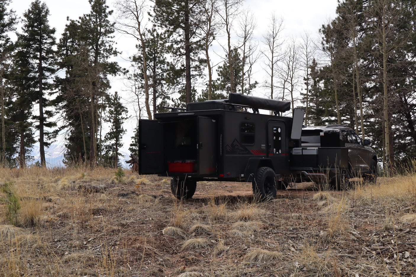 Overland trailer (RV rental) and SUV parked amidst pine and aspen trees, with mountains in the background, ready for adventure.