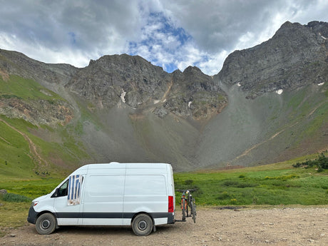 An RV rental camper van with bikes parked on a dirt road in a vast, rugged mountain landscape under cloudy skies.