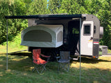 Grey rv rental camper with extended Thule awning, pop-out tent, and two chairs on grass amid green trees.