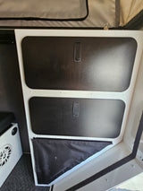 RV rental interior showing two black storage compartments with handles, a lower fabric pocket, and a speaker on a textured floor.
