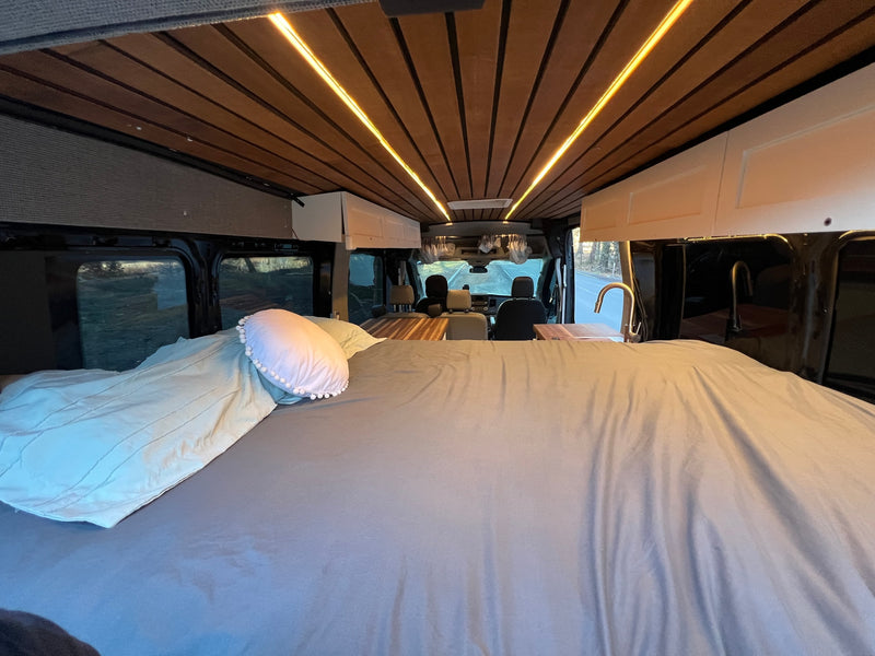 Cozy rv rental interior featuring a spacious bed, warm LED lighting on a wooden ceiling, and a kitchen area with a view of the road. — photo 16