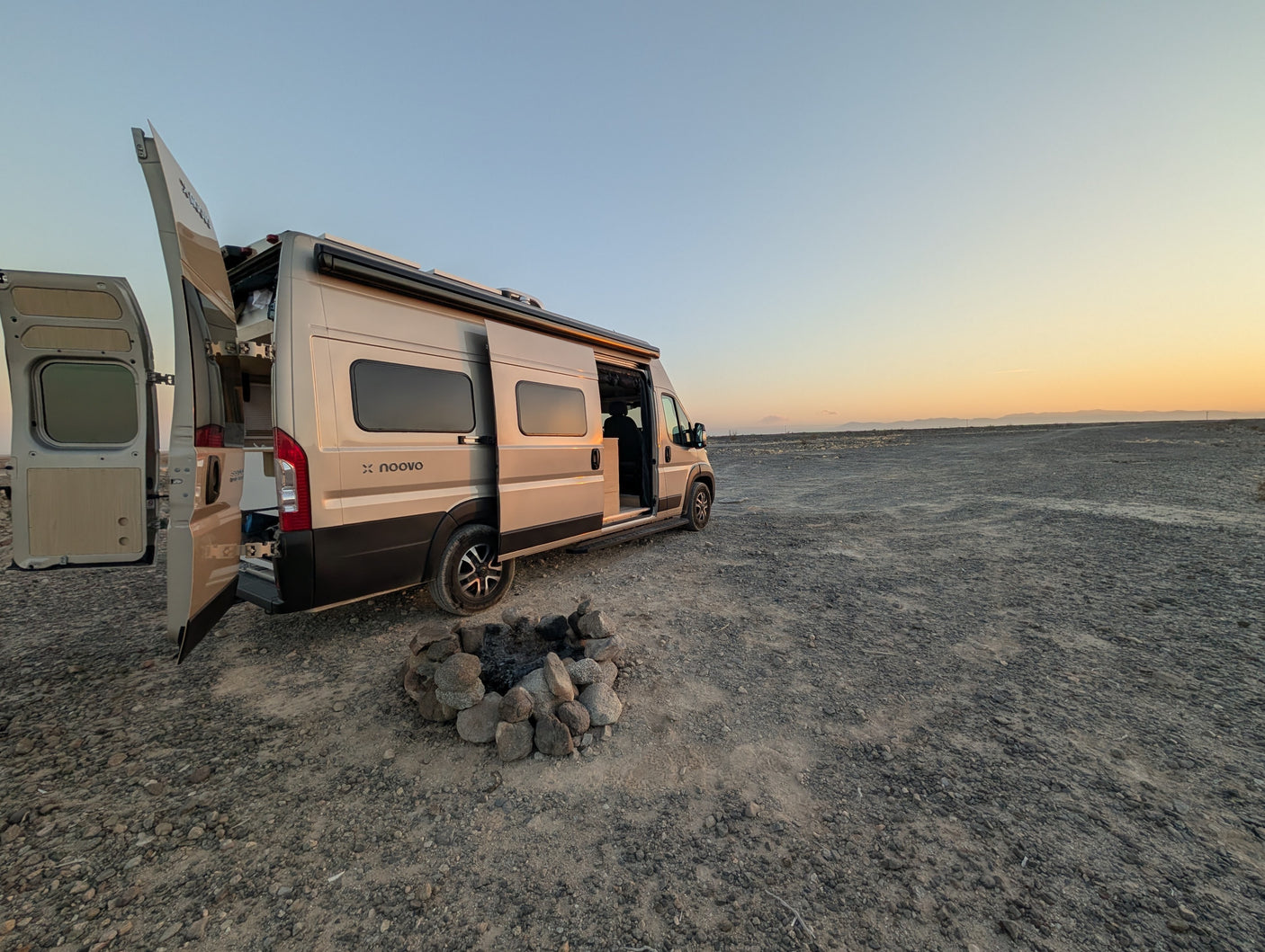 RV rental camper van with open doors, beside a rock fire pit on a gravel plain, under a fading sunset sky.