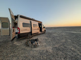 RV rental camper van with open doors, beside a rock fire pit on a gravel plain, under a fading sunset sky.