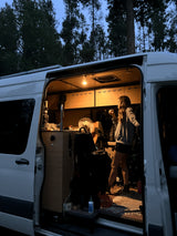 Brightly lit RV rental interior at dusk, with a person inside the cozy camper van among tall trees.