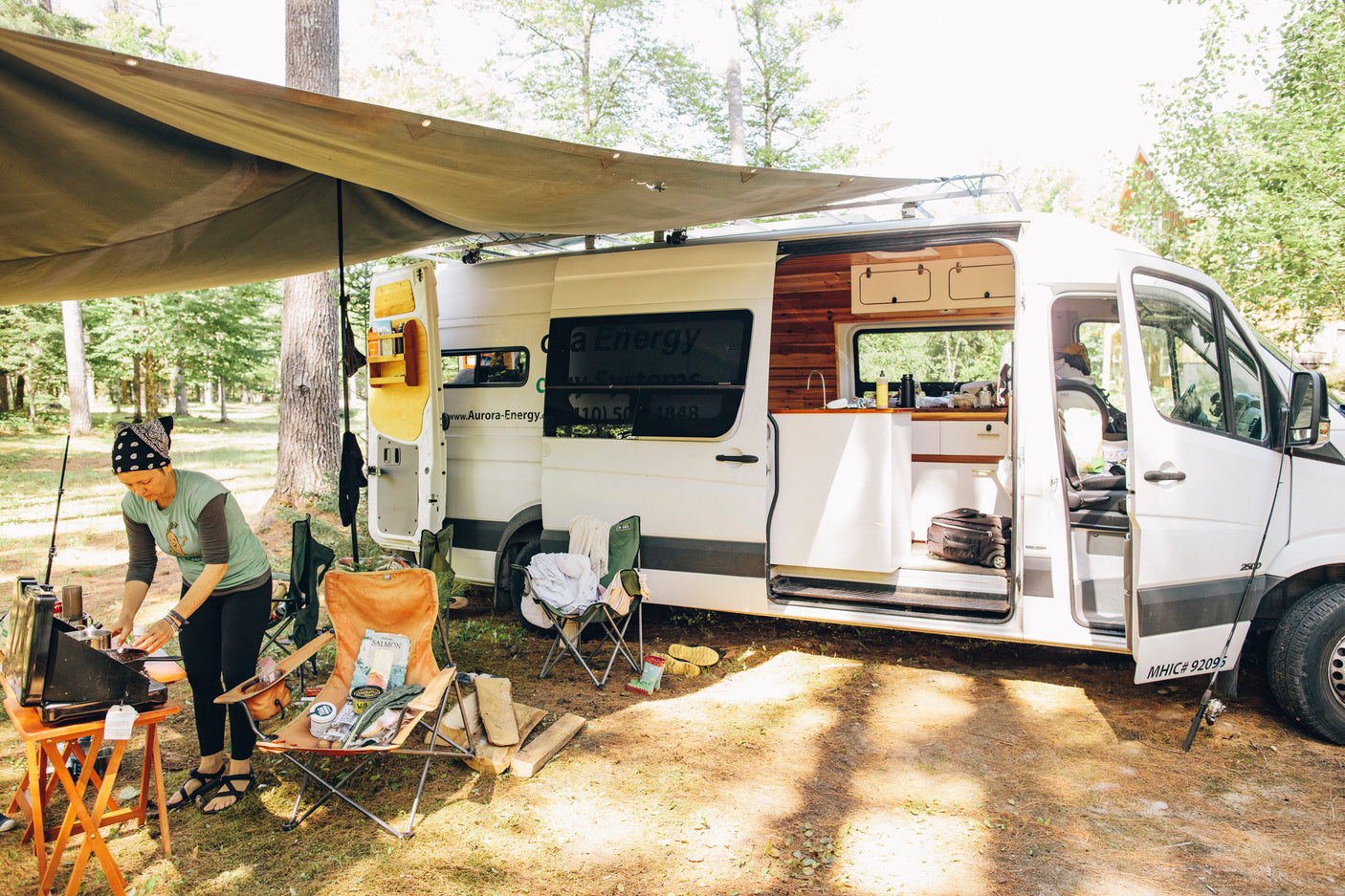 A woman cooks outside an RV rental camper van, revealing its cozy interior and campsite setup in a wooded area.
