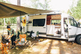 A woman cooks outside an RV rental camper van, revealing its cozy interior and campsite setup in a wooded area.