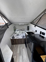 RV rental: Bright A-frame camper interior with a cozy bed, compact kitchen sink, and black mini-fridge on wood flooring.