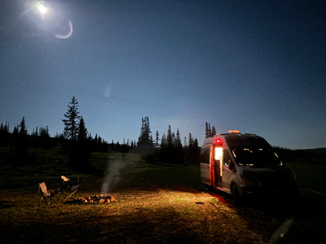 Night camping with an RV rental under a starry sky and bright moon. A camper van, campfire, and chairs complete the peaceful outdoor scene.