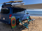 Enjoying Hawaii's coast with an rv rental. Blue pop-top camper van with surfboard, awning, and chairs on a sandy beach.