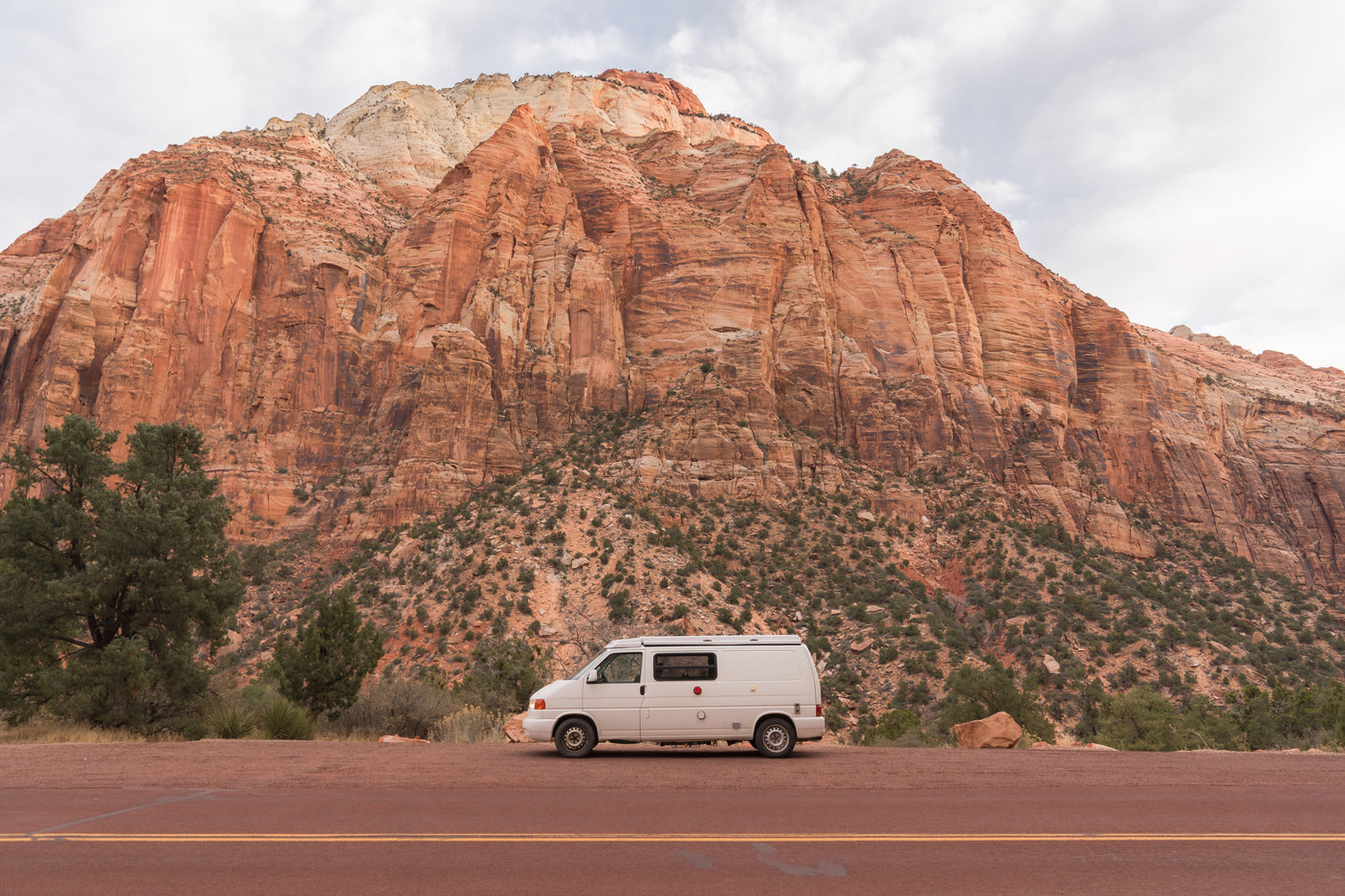 A white camper van, an RV rental, parked on a scenic road with towering red sandstone cliffs of Zion in the background.
