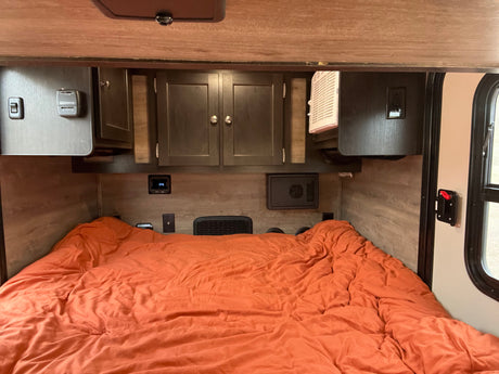 Cozy RV rental bedroom with an orange bed, dark wood cabinets, thermostat, power outlets, and a window.