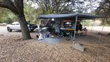 Explore the wild with our RV rental! A truck & off-road camper trailer, complete with outdoor kitchen and awning, set up at a leafy campsite.