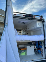 This RV rental's rear view reveals a comfy bed, white ceiling, storage, and exposed electrical system for off-grid power.