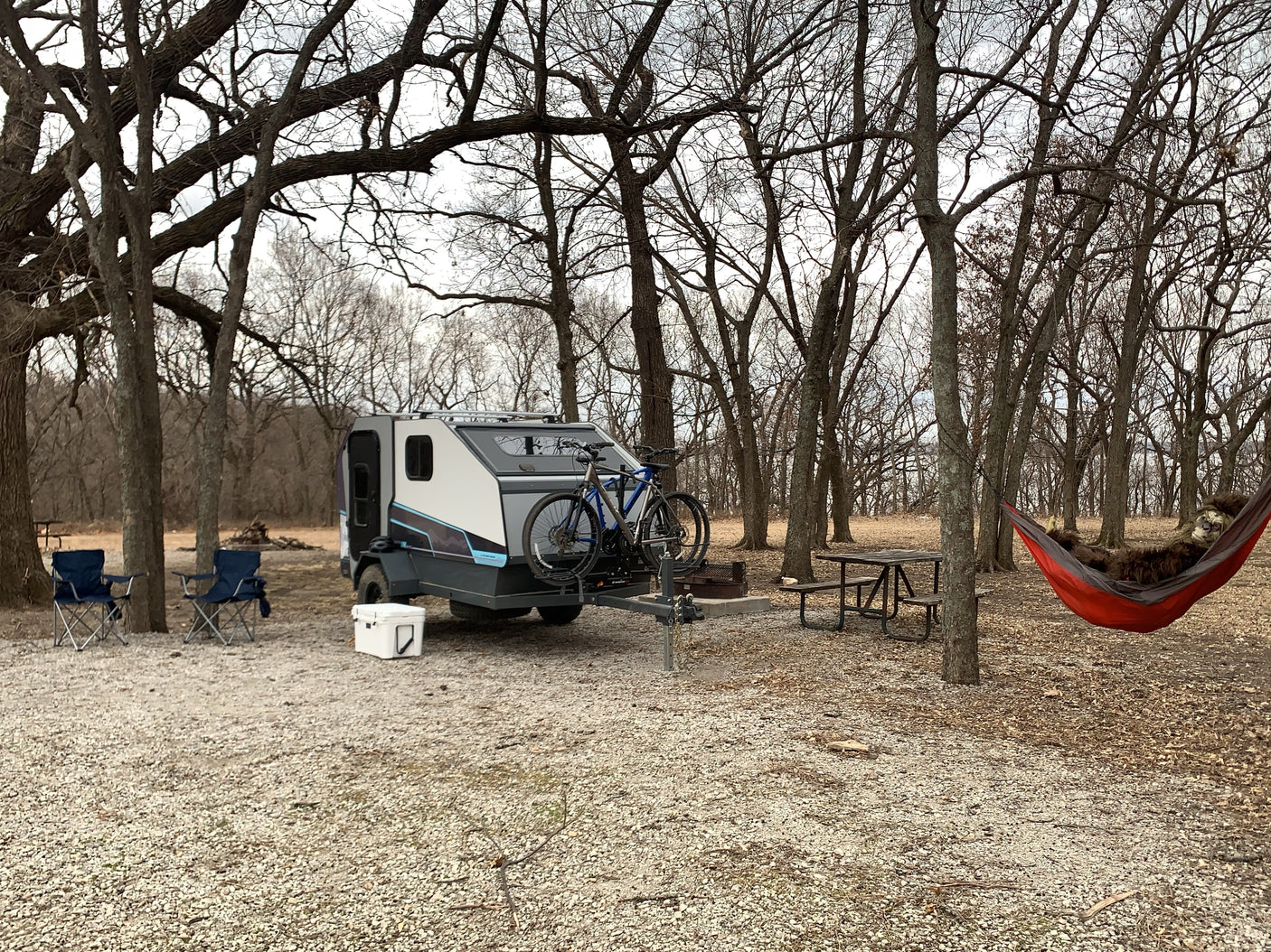 Teardrop camper (RV rental) & bikes at a campsite. Chairs, cooler, picnic table, and a relaxing Bigfoot in a hammock among bare trees.
