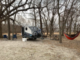 Teardrop camper (RV rental) & bikes at a campsite. Chairs, cooler, picnic table, and a relaxing Bigfoot in a hammock among bare trees.