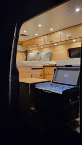 Cozy, well-lit interior of an rv rental camper van, featuring wooden walls/ceiling, a bed, cabinets, and an open portable fridge.