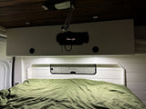 Inside an RV rental, a bed with a green duvet and a projector displaying 'Gilmore Girls' on overhead cabinets for cozy viewing.