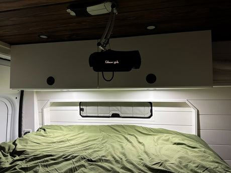 Inside an RV rental, a bed with a green duvet and a projector displaying 'Gilmore Girls' on overhead cabinets for cozy viewing.