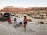 Woman walks by an RV rental camper van set up for desert camping with chairs and mountains in the background.
