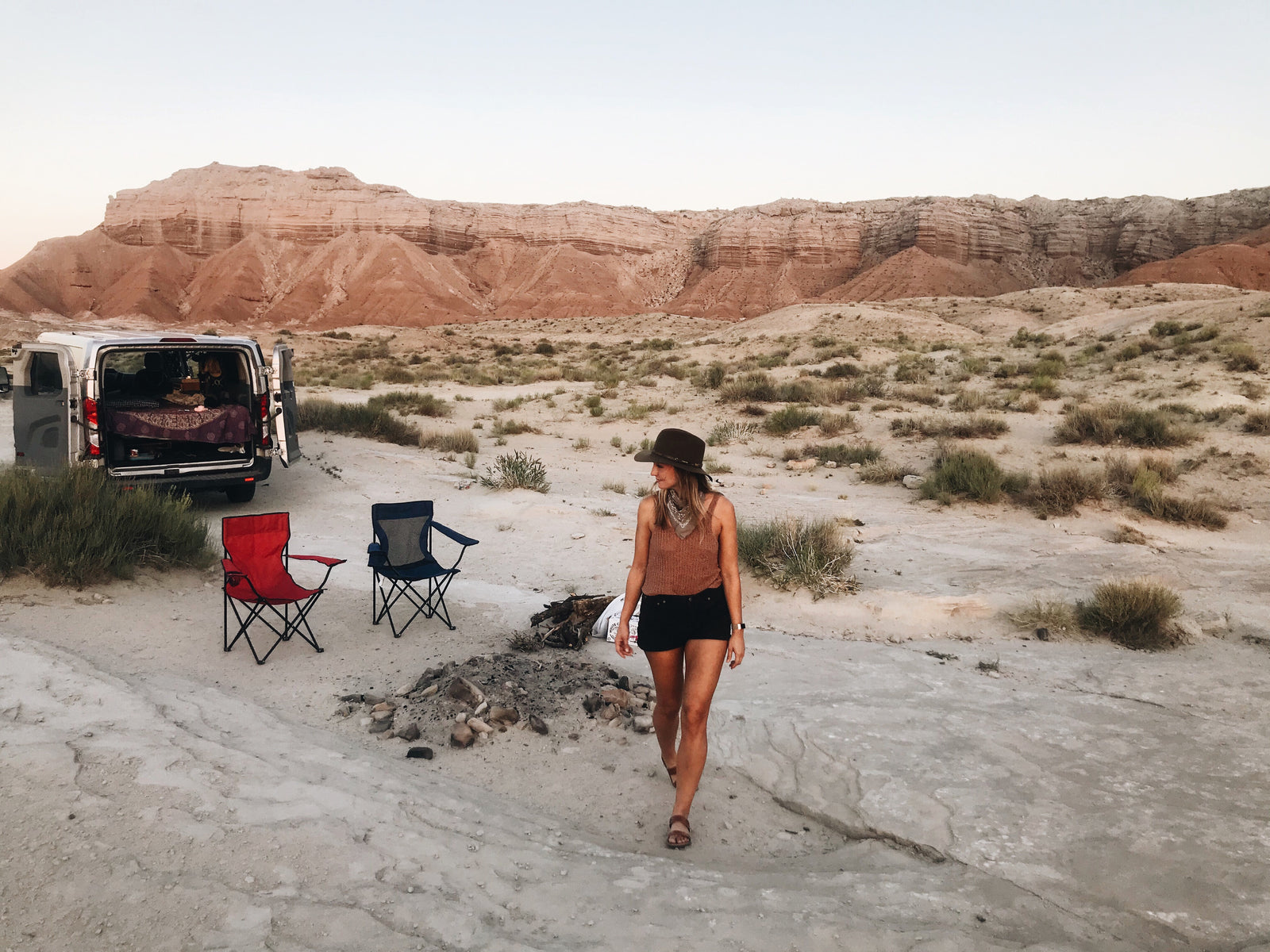 Woman walks by an RV rental camper van set up for desert camping with chairs and mountains in the background.