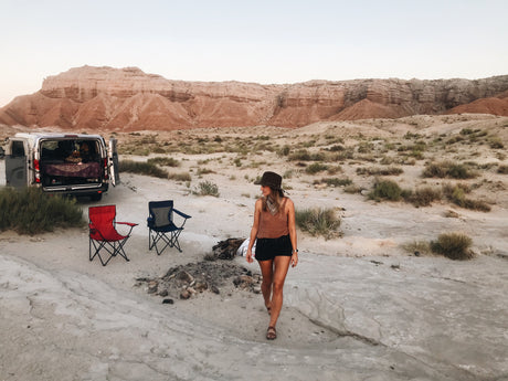 Woman walks by an RV rental camper van set up for desert camping with chairs and mountains in the background.