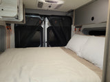 RV rental: Inside a camper van showing a cozy bed with white linens, black mesh screens on rear doors, overhead cabinets, and an AC unit.