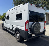 This white camper van, ready for adventure as an RV rental, features a rear spare tire and rooftop accessories for travel.