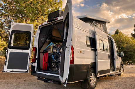 White Winnebago Solis rv rental parked with doors open, revealing a bike & gear. Features a pop-top roof for travel adventure.