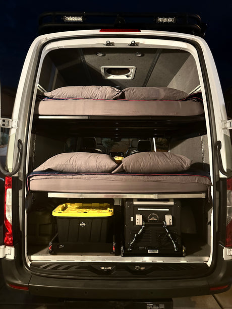 Inside an RV rental camper van: stacked bunk beds with bedding, storage bins, and a portable fridge/freezer for adventure.