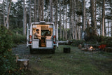 RV rental: A man works on his laptop inside a cozy van parked in a forest campsite with a crackling campfire.